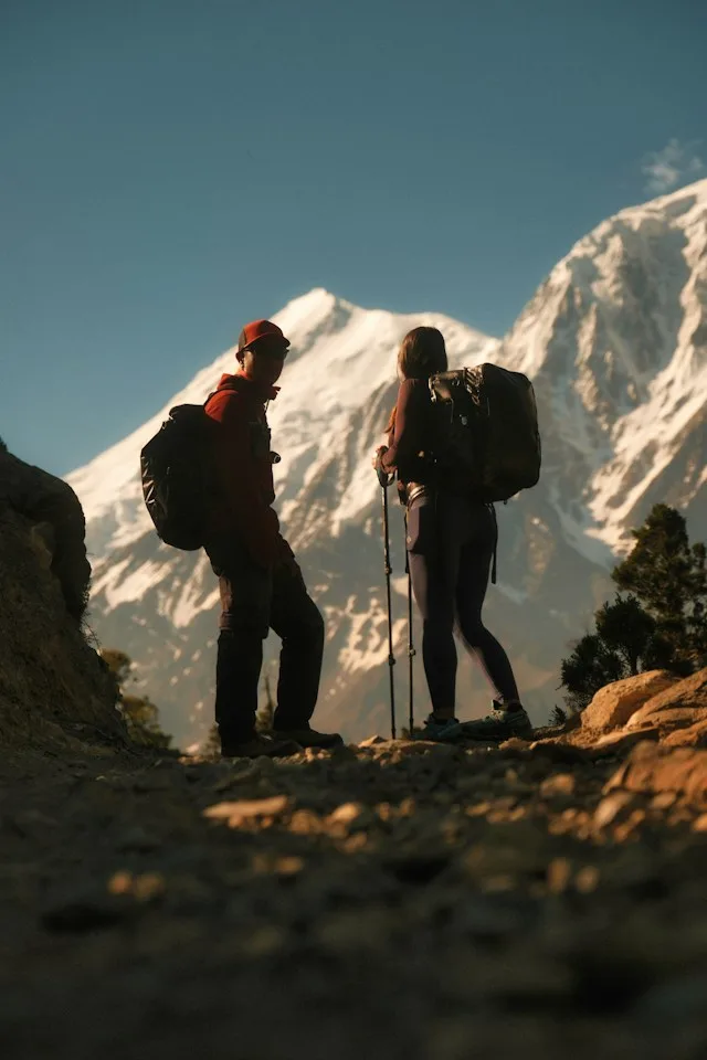 Two mountaineers with backpacks and trekking poles in front of a snow-covered peak